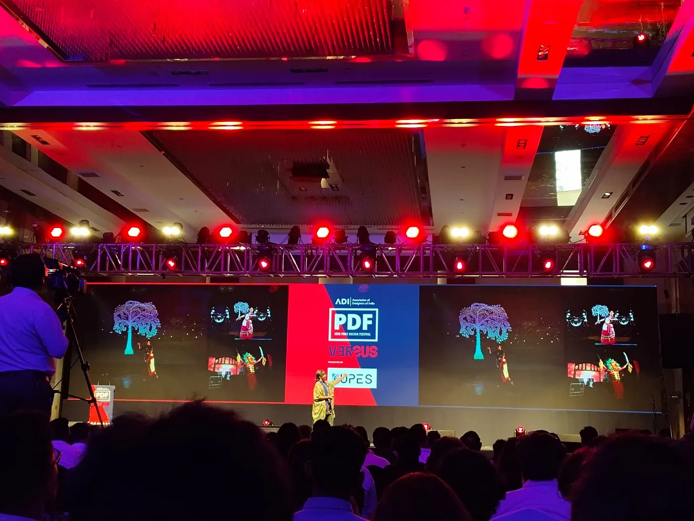 Upasana Nattoji Roy presenting on stage at the 18th Pune Design Festival — red and navy VERSUS backdrop, glowing projection tree and audience silhouettes