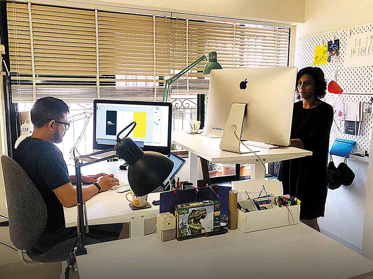 Two Switch & Roy designers in a bright studio — one seated at a large monitor, one standing at an iMac on a raised standing desk